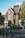 A charming brick house with a pitched roof and several chimneys, surrounded by greenery and set against a bright blue sky. The building features detailed masonry work and white-trimmed windows. It's located on a quiet street with black bollards lining the sidewalk.