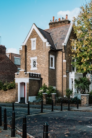 A charming brick house with a pitched roof and several chimneys, surrounded by greenery and set against a bright blue sky. The building features detailed masonry work and white-trimmed windows. It's located on a quiet street with black bollards lining the sidewalk.