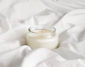 A jar candle with a clean, white surface sits nestled within soft, crumpled white fabric, creating a serene and calming atmosphere.