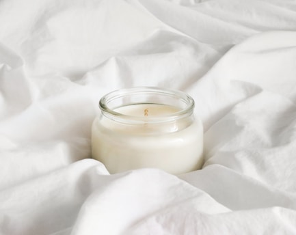 A jar candle with a clean, white surface sits nestled within soft, crumpled white fabric, creating a serene and calming atmosphere.