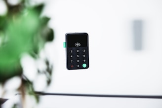 A digital payment card reader is floating in mid-air against a blurred background that includes green foliage and a gray wall. The device features a numeric keypad and has a contactless payment symbol displayed on its screen.
