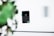 A digital payment card reader is floating in mid-air against a blurred background that includes green foliage and a gray wall. The device features a numeric keypad and has a contactless payment symbol displayed on its screen.