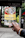 A hand holding a colorful brochure or pamphlet with images and text, possibly related to a cultural or historical site. The background is blurred, showing some greenery and a dark sign with faded text.