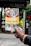 A hand holding a colorful brochure or pamphlet with images and text, possibly related to a cultural or historical site. The background is blurred, showing some greenery and a dark sign with faded text.