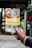 A hand holding a colorful brochure or pamphlet with images and text, possibly related to a cultural or historical site. The background is blurred, showing some greenery and a dark sign with faded text.