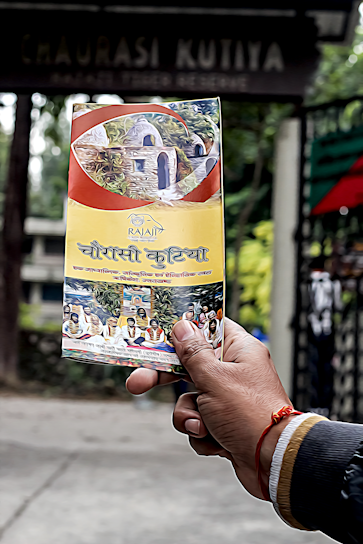 A hand holding a colorful brochure or pamphlet with images and text, possibly related to a cultural or historical site. The background is blurred, showing some greenery and a dark sign with faded text.