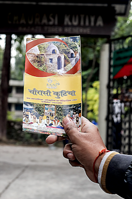 A hand holding a colorful brochure or pamphlet with images and text, possibly related to a cultural or historical site. The background is blurred, showing some greenery and a dark sign with faded text.