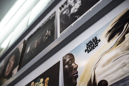 An angled view of a series of vinyl record covers displayed on a shelf, featuring a prominently visible cover with an abstract portrait and text. The colors are muted and the lighting is soft, creating a vintage atmosphere.