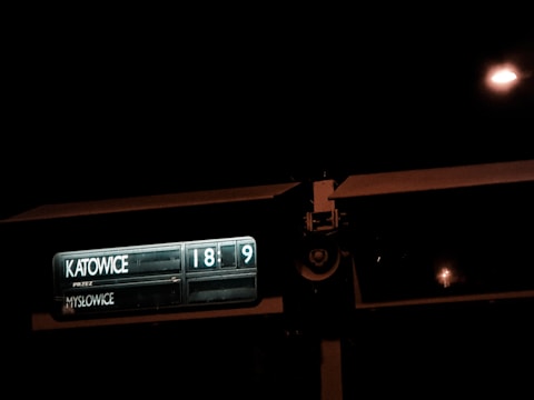 A dimly lit train or bus sign displaying the destinations 'Katowice' and 'Mysłowice' with digital numbers indicating the next arrival or departure times, set against a dark night sky.