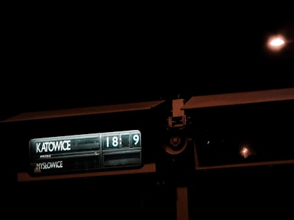 A dimly lit train or bus sign displaying the destinations 'Katowice' and 'Mysłowice' with digital numbers indicating the next arrival or departure times, set against a dark night sky.
