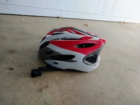 A cycling helmet with red, white, and black designs is placed on a concrete floor. The helmet has several ventilation holes and a clasp is attached for securing it.