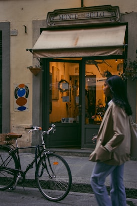 A cozy street scene featuring an entrance to a quaint shop named Monte Bello. The shopfront has a warm glow emanating from inside, suggesting a welcoming ambiance. Outside, a bicycle is parked casually near the entrance, and a person wearing a tan coat is walking past, partially in motion.