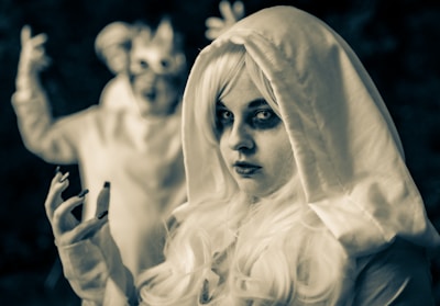 A person dressed in a costume with a hooded cloak and striking makeup, featuring intense eye makeup and dark circles. The blurred figure in the background shares similar attire, creating an eerie and mysterious atmosphere.