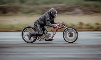 A person dressed in black leather riding gear is speeding on a minimalist, custom-built motorcycle with a prominent red frame and extended handlebars. The setting appears to be an outdoor track, with a blurred background suggesting motion at high speed.
