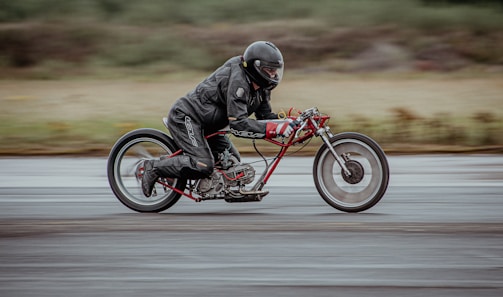 A person dressed in black leather riding gear is speeding on a minimalist, custom-built motorcycle with a prominent red frame and extended handlebars. The setting appears to be an outdoor track, with a blurred background suggesting motion at high speed.
