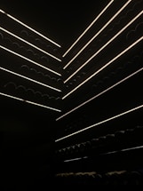 Several rows of wine bottles are stacked horizontally in a dimly lit storage area with narrow strips of light illuminating the shelves. The scene creates a sharp contrast between the dark environment and the subtle, glowing lines that highlight the outlines of the bottles.