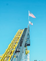 A steep roller coaster track painted in bright yellow and blue ascends towards the sky. At the peak, two flags are raised on tall poles: the American flag and a California state flag. The background is a clear, bright blue sky.
