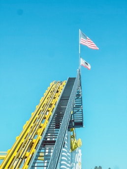 A steep roller coaster track painted in bright yellow and blue ascends towards the sky. At the peak, two flags are raised on tall poles: the American flag and a California state flag. The background is a clear, bright blue sky.