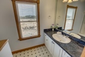 A spacious bathroom with a large window featuring wooden frames overlooking an outdoor view. The interior includes a black marble countertop with a single sink, surrounded by white cabinets. There is a mirror above the sink, and the floor has a white and black checkered pattern. Two small vases with flowers are placed on the counter, adding a decorative element.