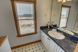 A spacious bathroom with a large window featuring wooden frames overlooking an outdoor view. The interior includes a black marble countertop with a single sink, surrounded by white cabinets. There is a mirror above the sink, and the floor has a white and black checkered pattern. Two small vases with flowers are placed on the counter, adding a decorative element.