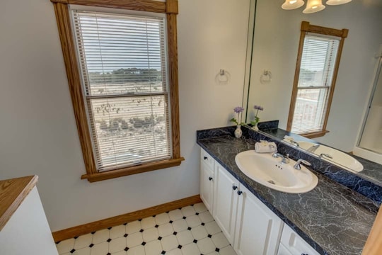 A spacious bathroom with a large window featuring wooden frames overlooking an outdoor view. The interior includes a black marble countertop with a single sink, surrounded by white cabinets. There is a mirror above the sink, and the floor has a white and black checkered pattern. Two small vases with flowers are placed on the counter, adding a decorative element.