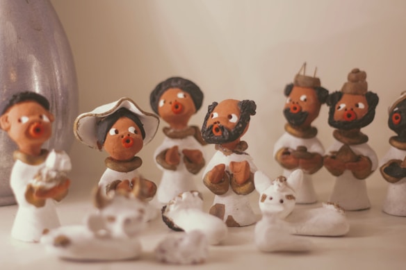 Clay figurines are arranged on a surface, depicting people in traditional attire. The figures display expressive faces with distinct eyes and mouths, showcasing various poses and gestures. They are surrounded by small white animal figurines, creating a lively and cultural scene.