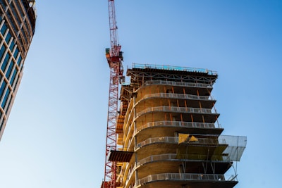 A partially constructed building with multiple visible floors and a prominent red construction crane adjacent to it. The structure appears to be modern, with metal railings on its edges. A nearby completed building with a grid-like window pattern is also present.