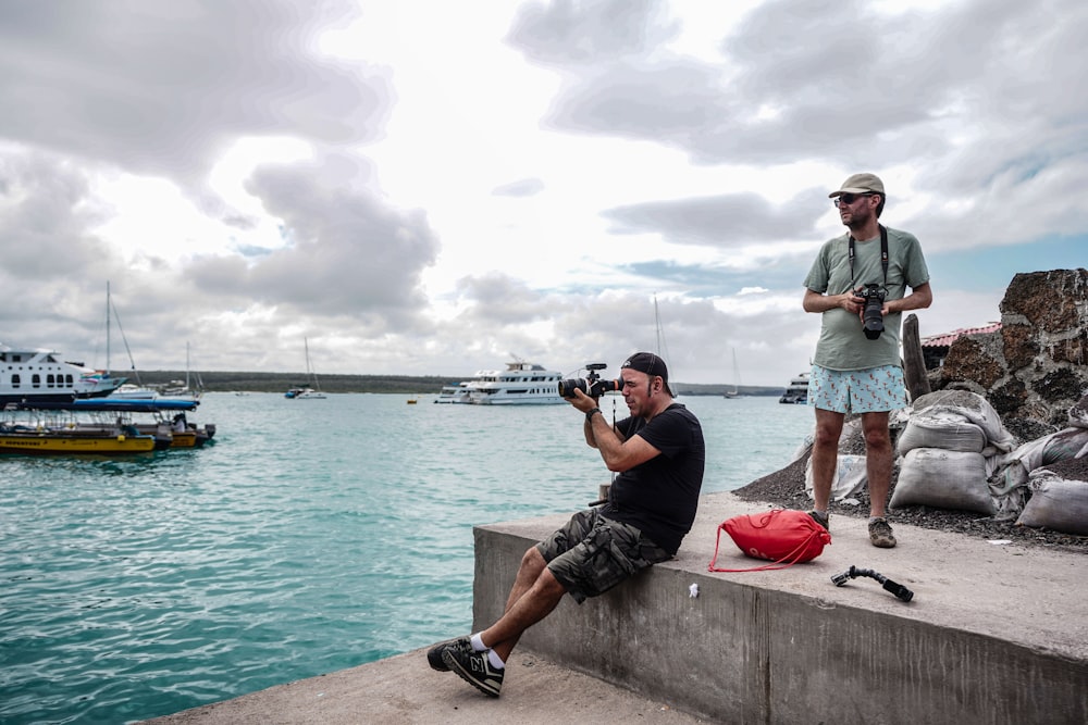 un homme assis sur un quai avec une canne à pêche et un homme debout sur un rocher avec des bateaux