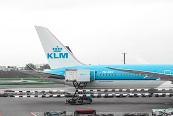 A section of a KLM airplane is parked on the tarmac. The tail and rear part of the fuselage are visible with the KLM logo prominently displayed. In the foreground, there is ground equipment and luggage carts.