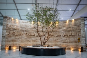 A large, elegantly designed interior space features a tall, live tree planted in a circular black planter. The background is a marble-like textured wall with ornate Arabic script illuminated by soft spotlights from above. The ceiling is high with structural beams visible.
