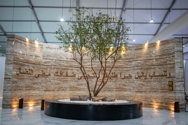 A large, elegantly designed interior space features a tall, live tree planted in a circular black planter. The background is a marble-like textured wall with ornate Arabic script illuminated by soft spotlights from above. The ceiling is high with structural beams visible.