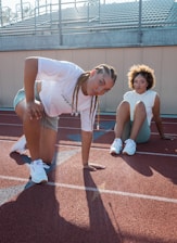 Two individuals in athletic attire are positioned on a running track. One is kneeling with one hand on the ground, while the other is seated with legs bent. Both are making direct eye contact with the camera. The setting suggests a sports field, with bleachers visible in the background.