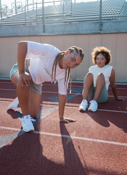 Two individuals in athletic attire are positioned on a running track. One is kneeling with one hand on the ground, while the other is seated with legs bent. Both are making direct eye contact with the camera. The setting suggests a sports field, with bleachers visible in the background.