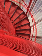 A spiral staircase with bright red railing and steps, contrasted with a steel mesh that allows light and shadows to play on the surfaces. The perspective offers a downward view, accentuating the curvature and symmetry of the structure.