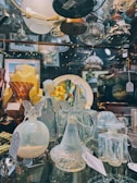 A glass display case filled with various elegant glassware items including vases, bottles, and decorative pieces featuring intricate designs. Many of the objects have price tags attached, suggesting they are for sale. The background shows additional glass items and decorative lights that add a warm ambiance.