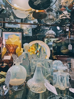 A glass display case filled with various elegant glassware items including vases, bottles, and decorative pieces featuring intricate designs. Many of the objects have price tags attached, suggesting they are for sale. The background shows additional glass items and decorative lights that add a warm ambiance.