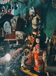A collection of Japanese cultural items and artifacts are displayed on shelves in a dimly lit room. Prominent among them is a doll dressed in a traditional kimono with floral patterns in red and white. Above the doll, various masks, including a kitsune and oni mask, are hung on the wall. Other items in the display include fans, teapots, baskets, and framed artwork. The ambient light casts shadows, creating a cozy and nostalgic atmosphere.