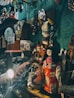 A collection of Japanese cultural items and artifacts are displayed on shelves in a dimly lit room. Prominent among them is a doll dressed in a traditional kimono with floral patterns in red and white. Above the doll, various masks, including a kitsune and oni mask, are hung on the wall. Other items in the display include fans, teapots, baskets, and framed artwork. The ambient light casts shadows, creating a cozy and nostalgic atmosphere.