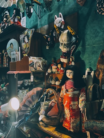 A collection of Japanese cultural items and artifacts are displayed on shelves in a dimly lit room. Prominent among them is a doll dressed in a traditional kimono with floral patterns in red and white. Above the doll, various masks, including a kitsune and oni mask, are hung on the wall. Other items in the display include fans, teapots, baskets, and framed artwork. The ambient light casts shadows, creating a cozy and nostalgic atmosphere.