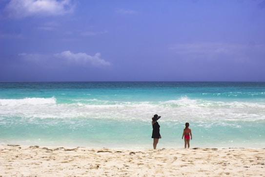 A woman wearing a black dress and large hat stands on a sandy beach, looking out at the ocean alongside a child in a pink swimsuit. The clear blue sky and turquoise water create a serene, peaceful setting.