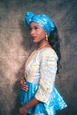 A person wearing a vibrant traditional outfit consisting of a blue patterned top with yellow details and a matching blue headwrap. The person has long black hair, accessorized with gold earrings and a gold necklace. They are posing sideways with a subtle smile against a textured background.