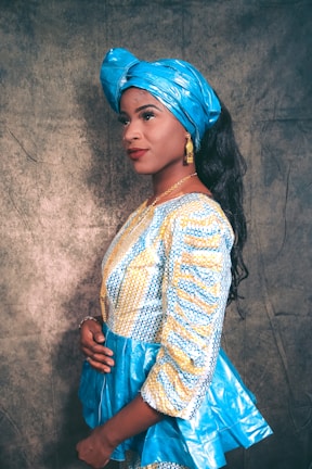 A person wearing a vibrant traditional outfit consisting of a blue patterned top with yellow details and a matching blue headwrap. The person has long black hair, accessorized with gold earrings and a gold necklace. They are posing sideways with a subtle smile against a textured background.
