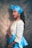 A person wearing a vibrant traditional outfit consisting of a blue patterned top with yellow details and a matching blue headwrap. The person has long black hair, accessorized with gold earrings and a gold necklace. They are posing sideways with a subtle smile against a textured background.