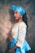 A person wearing a vibrant traditional outfit consisting of a blue patterned top with yellow details and a matching blue headwrap. The person has long black hair, accessorized with gold earrings and a gold necklace. They are posing sideways with a subtle smile against a textured background.