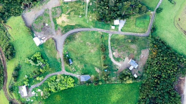 Aerial view of a rural area with a mix of fields, trees, and several small buildings connected by dirt roads. The landscape is predominantly green with patches of grass and forested areas.