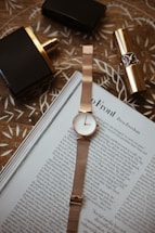 A rose gold wristwatch is neatly placed on top of an open magazine with visible text. Nearby, there's a black and gold perfume bottle and a gold lipstick tube on a patterned wooden surface.