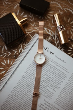 A rose gold wristwatch is neatly placed on top of an open magazine with visible text. Nearby, there's a black and gold perfume bottle and a gold lipstick tube on a patterned wooden surface.