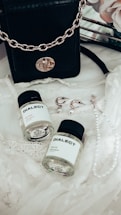 A black leather handbag with a gold chain sits on a delicate lace fabric. Nearby, there are two small bottles labeled 'DIALECT.' Earrings with pearl accents and a pearl necklace are also arranged on the lace.