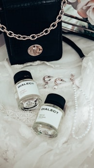 A black leather handbag with a gold chain sits on a delicate lace fabric. Nearby, there are two small bottles labeled 'DIALECT.' Earrings with pearl accents and a pearl necklace are also arranged on the lace.