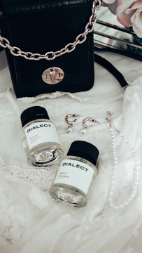 A black leather handbag with a gold chain sits on a delicate lace fabric. Nearby, there are two small bottles labeled 'DIALECT.' Earrings with pearl accents and a pearl necklace are also arranged on the lace.